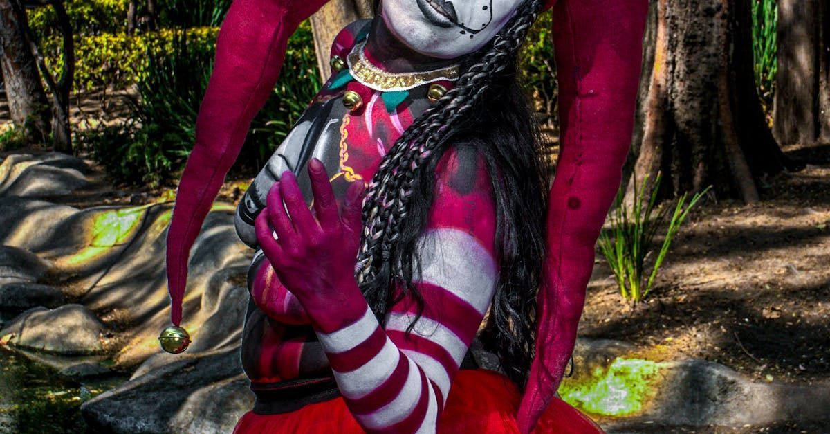 Person in jester body paint, pink and white stripes, in a park, arms crossed.