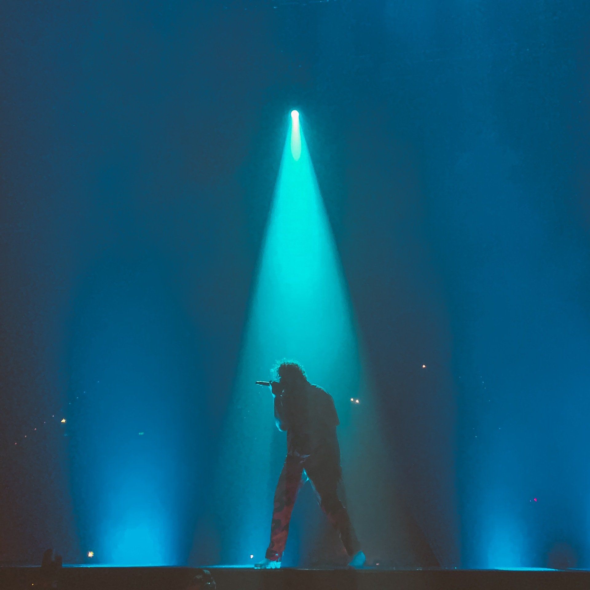 A figure on stage in a bright blue spotlight, performing with a microphone.
