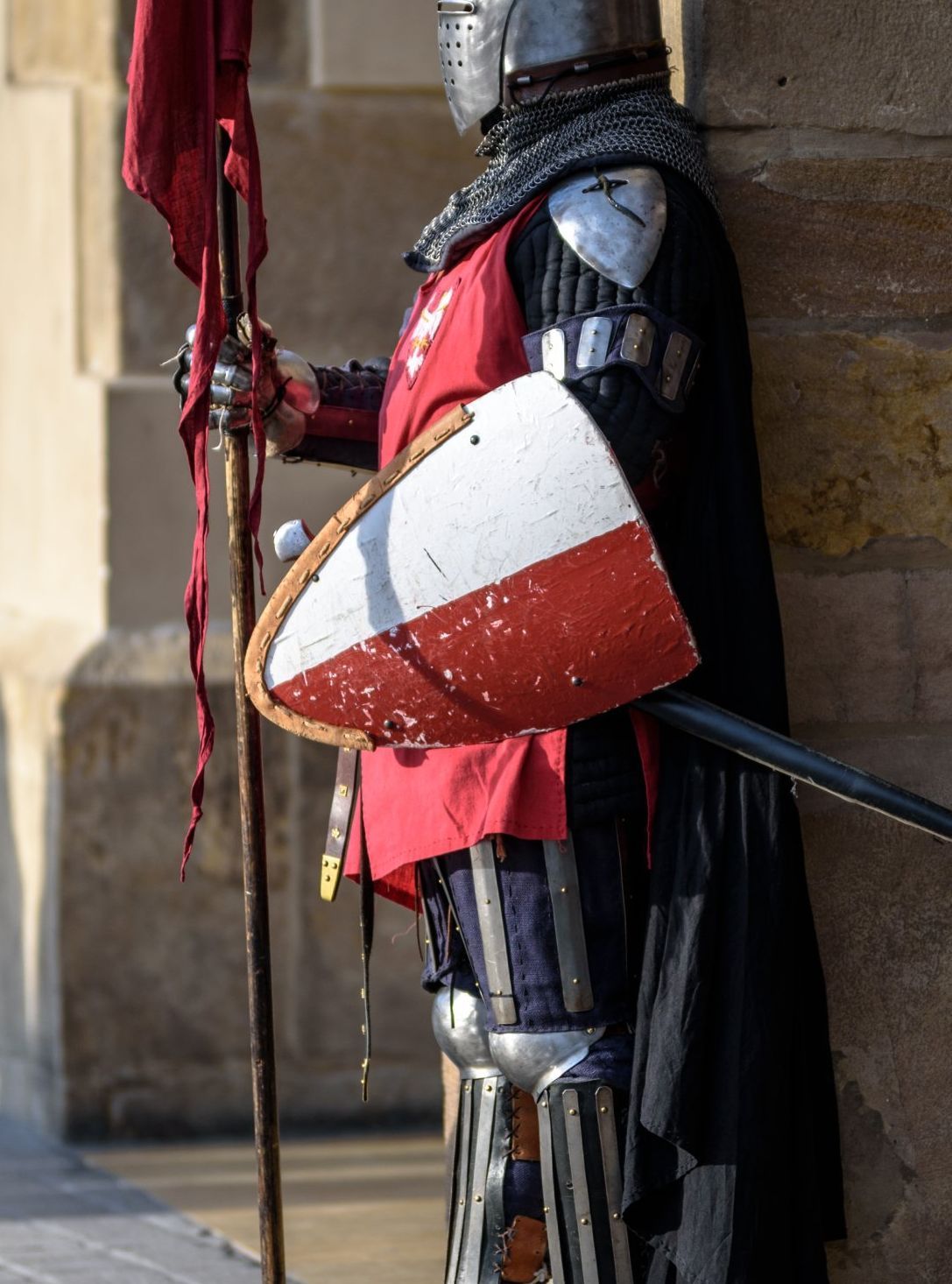 Knight in armor holding a shield and banner, leaning against a stone wall. Red and white shield.