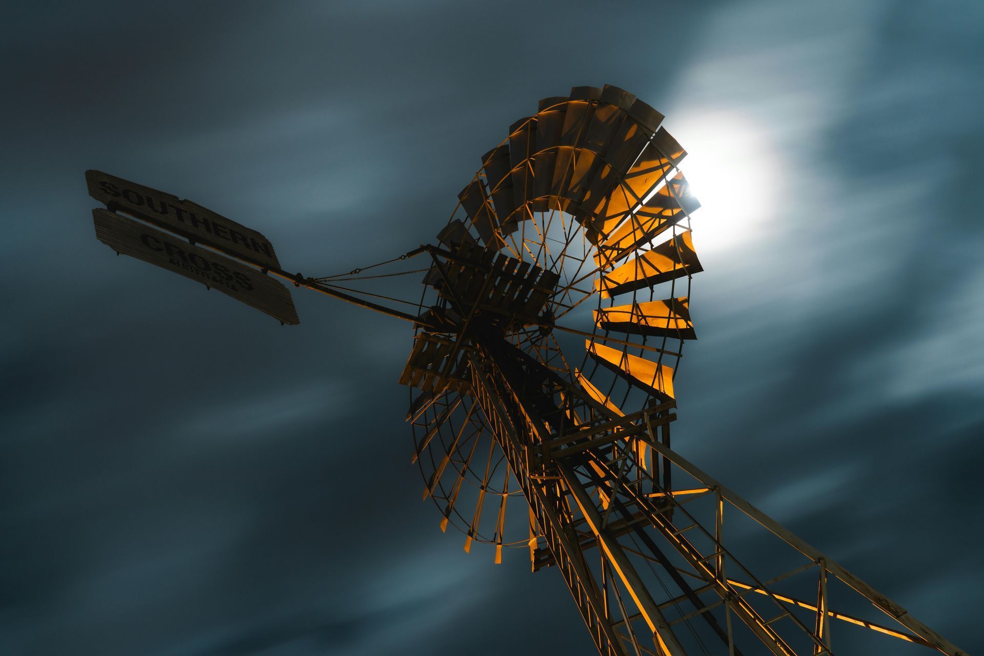 Windmill against a dark, cloudy sky, sun partially visible behind the blades.