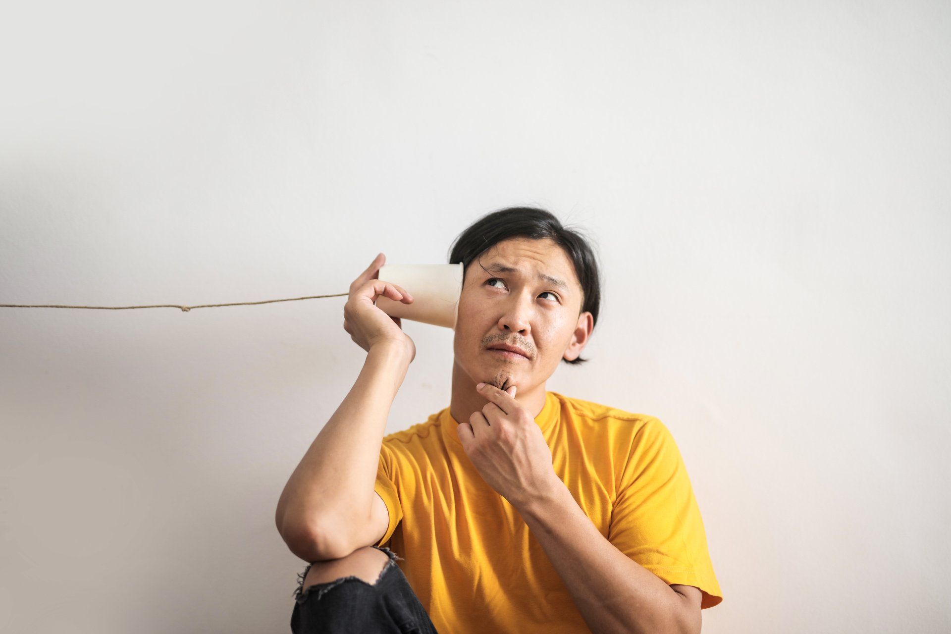 Man listening through a paper cup string telephone, looking upward with a thoughtful expression.