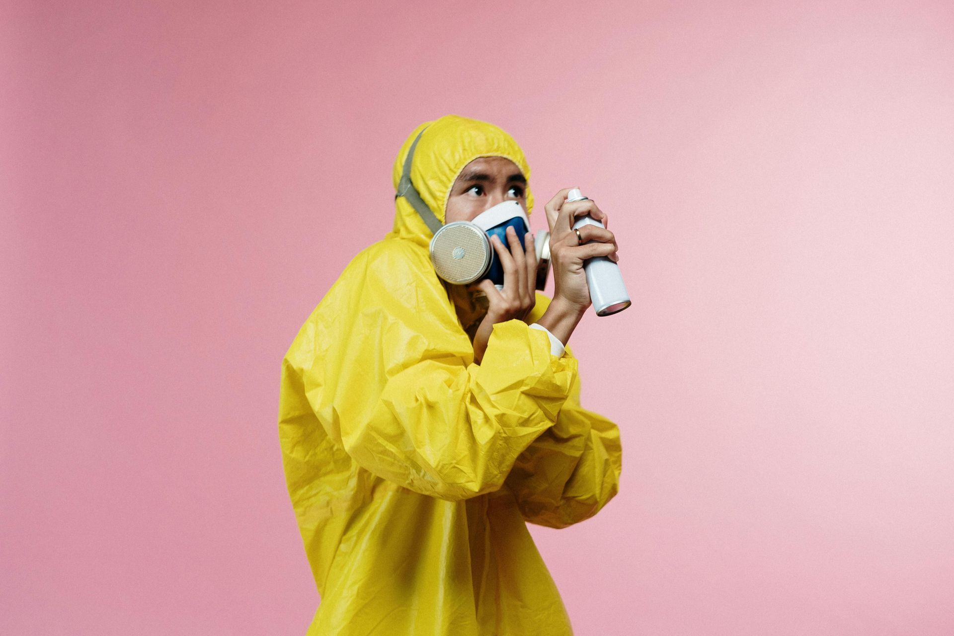 Person in yellow hazmat suit and respirator holds spray can, looking away, against pink background.