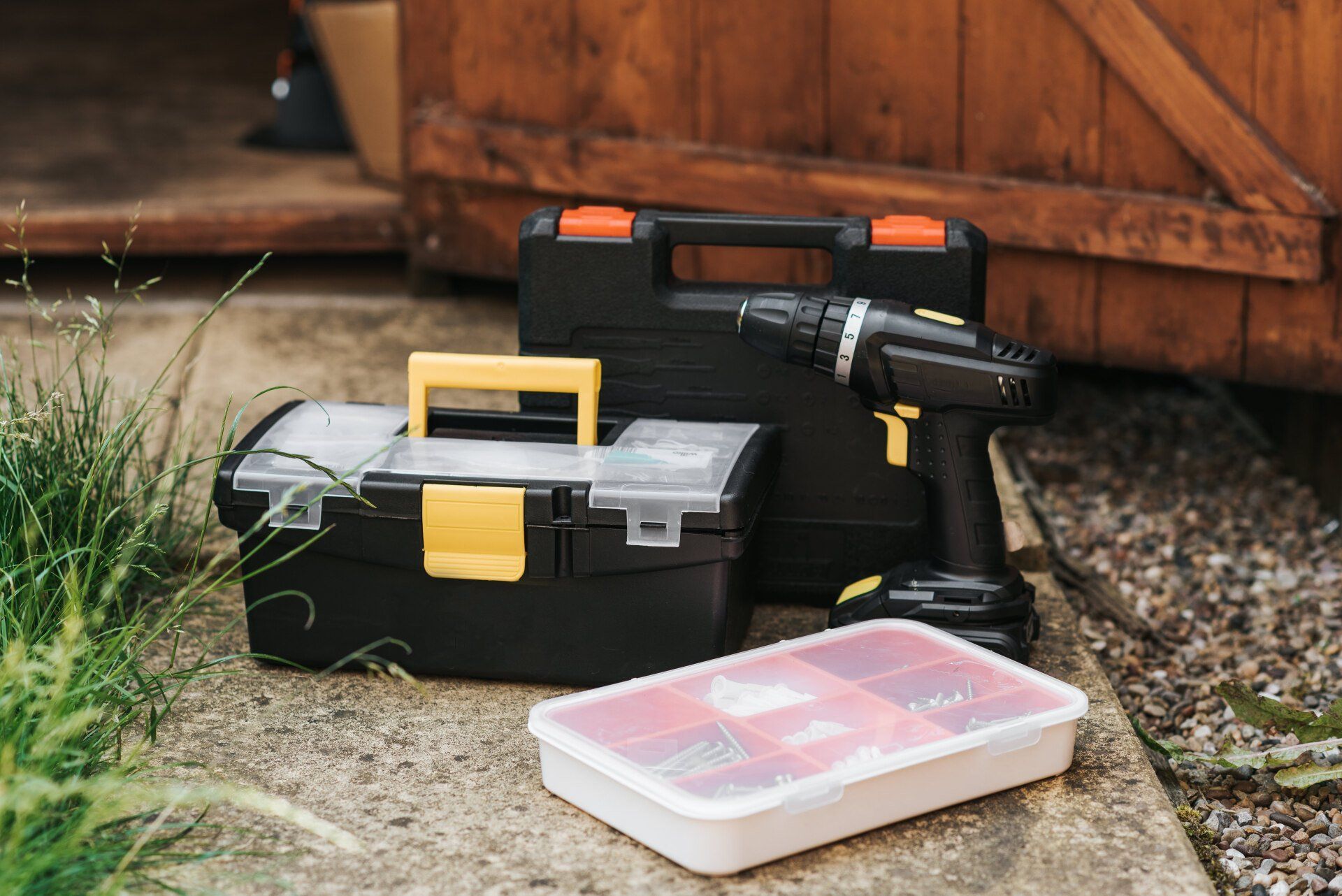 Toolboxes, cordless drill, and parts container on concrete surface near a wooden shed.
