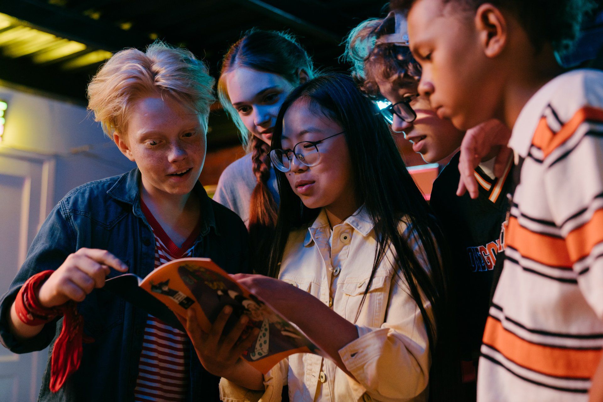 Group of kids looking at a book, some smiling. Indoors, dim lighting.