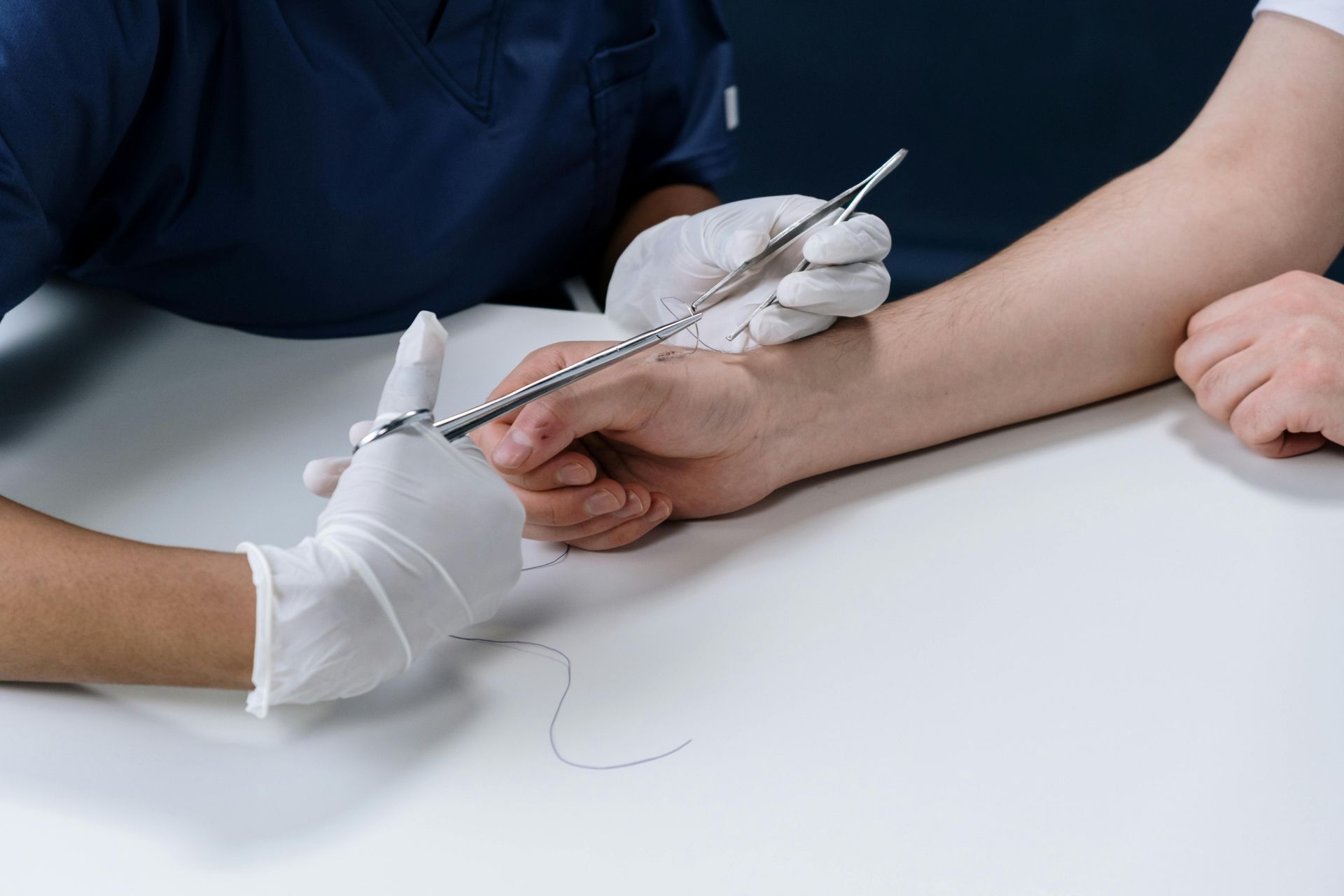 Person's wrist being held as gloved hand uses surgical instrument to remove a dark hair on a white table.
