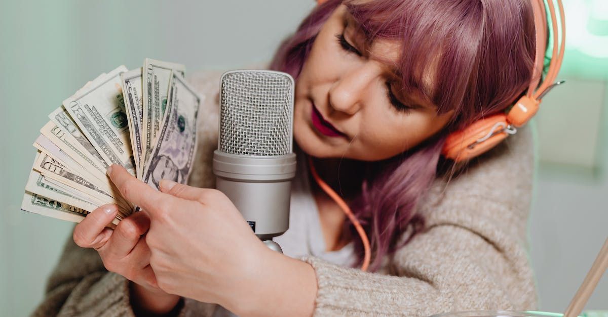 Woman with purple hair leans into a microphone, holding a fan of cash while wearing headphones.