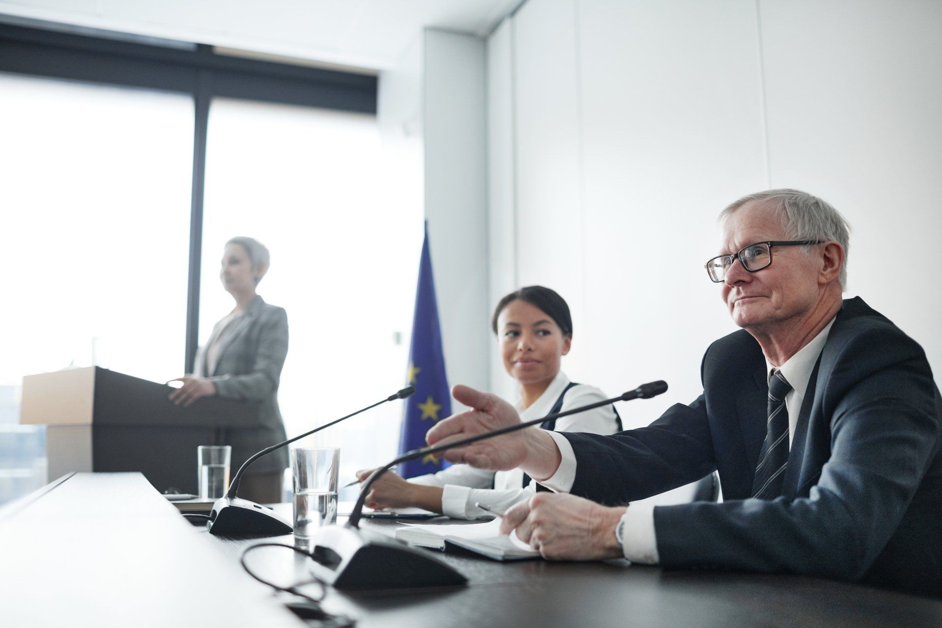 Man in suit gestures at a table during a press conference. Another woman and a speaker stand nearby.