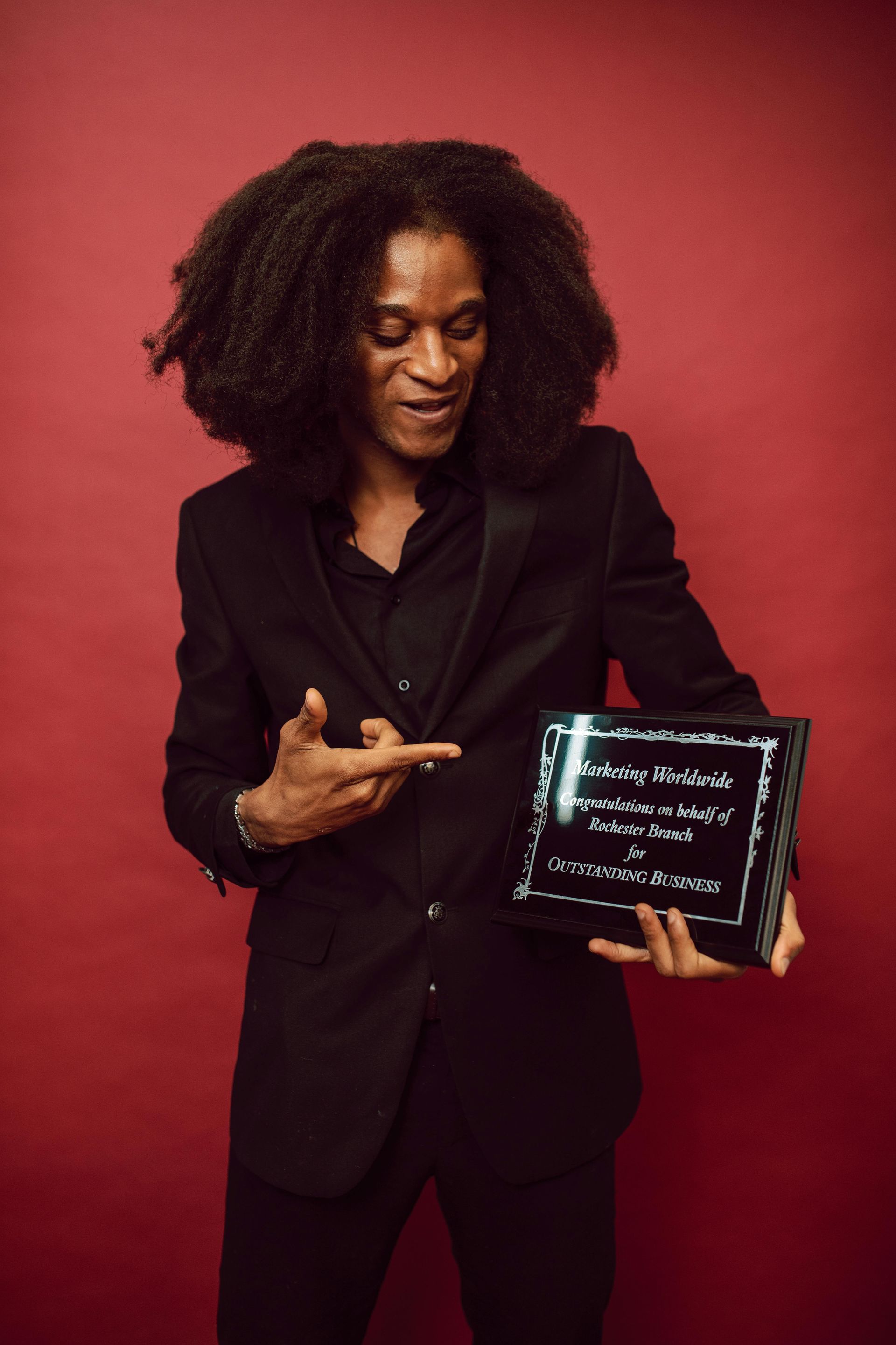 Person in black suit points at an award box against a red backdrop. The award box is dark with white text.
