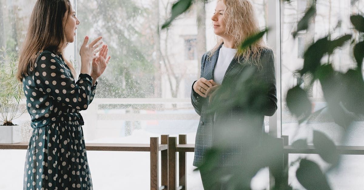 Two women talking near a window; one gesturing. Plants in the foreground.
