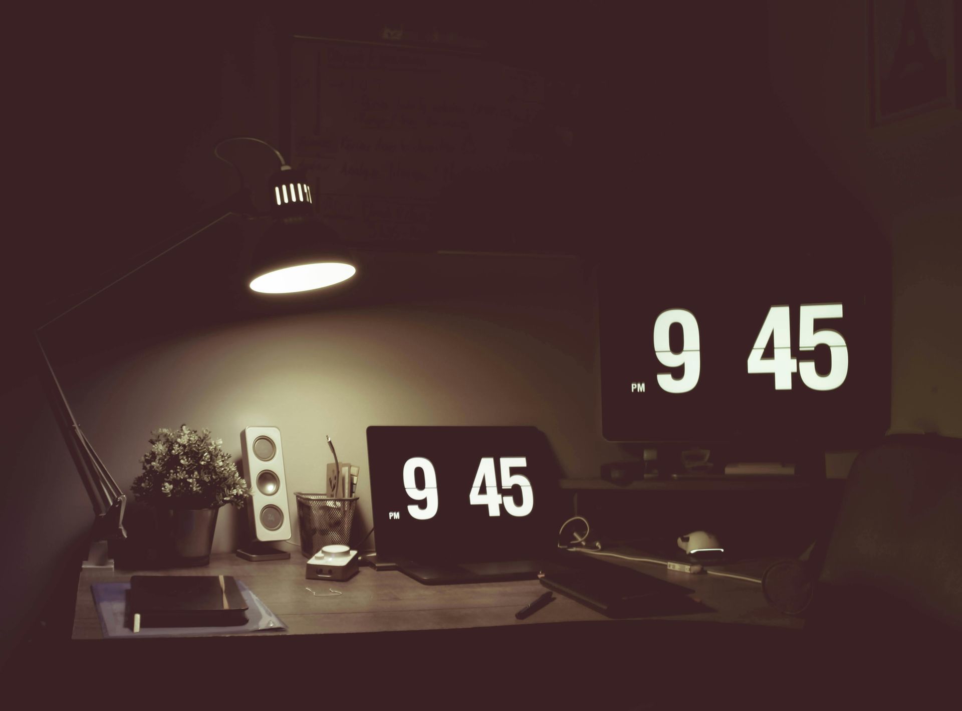 Desk with two clocks displaying 9:45, a lamp, speakers, and a plant in a dimly lit room.