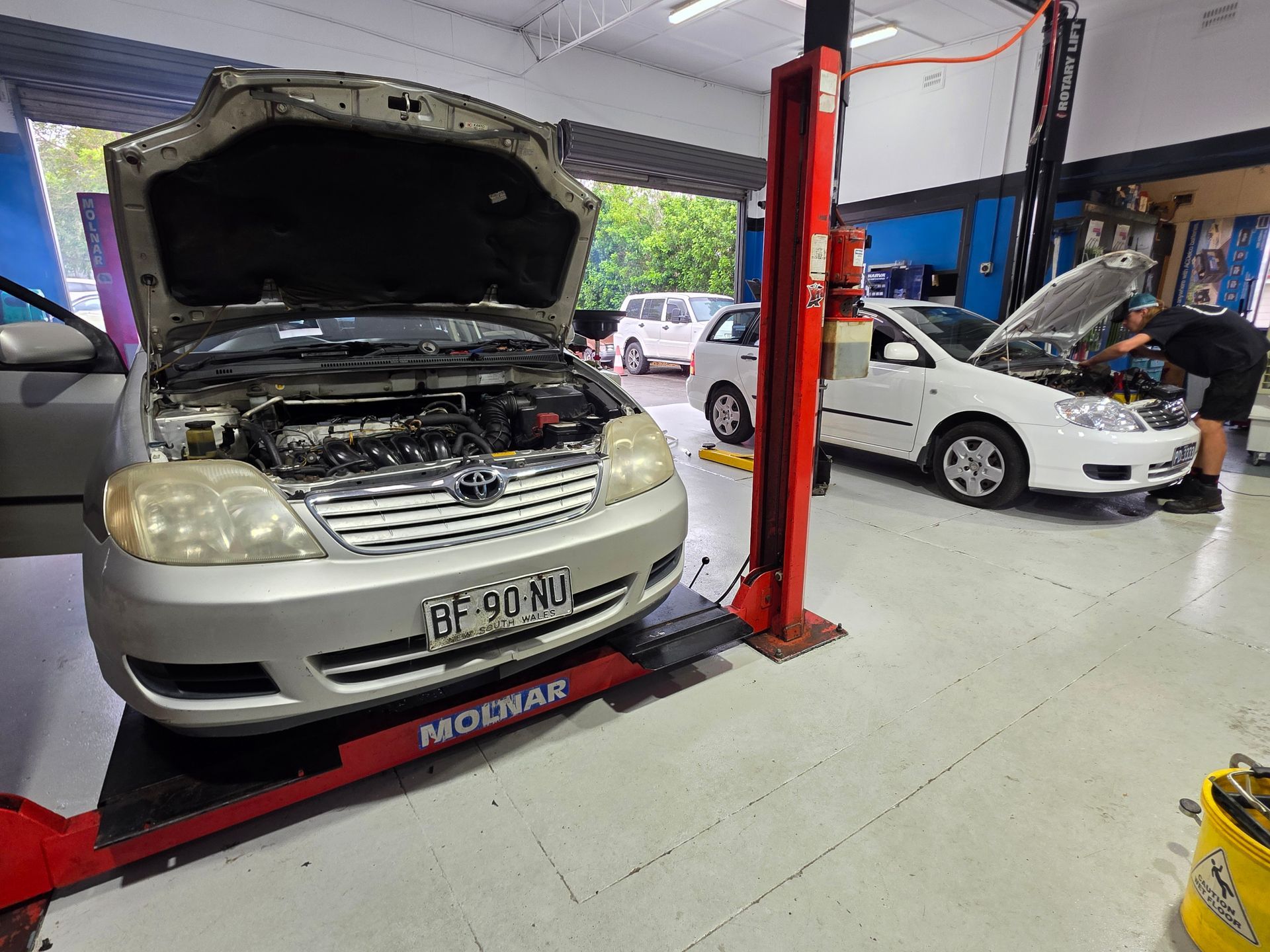 Cars in an auto repair shop, with one silver car raised on a lift and its hood open. — M T Automotive In Wallsend, NSW