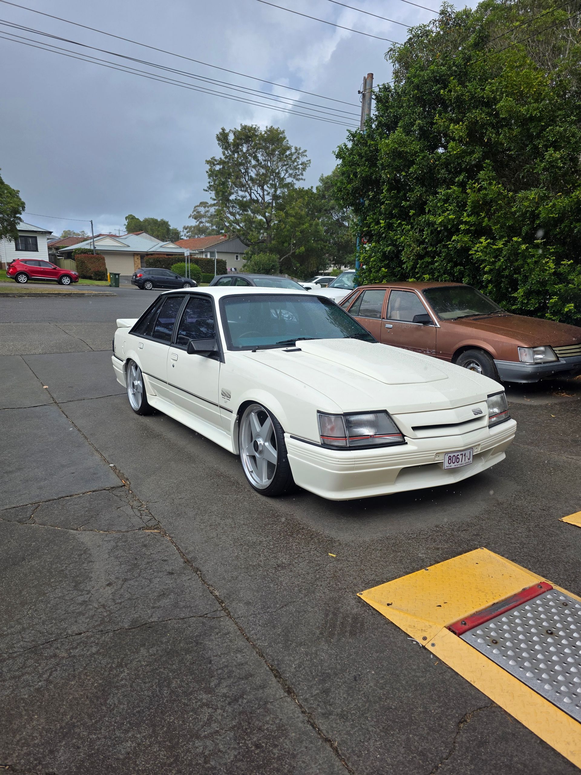 White sports car parked in a wet lot beside a brown car under a cloudy sky. — M T Automotive In Wallsend, NSW