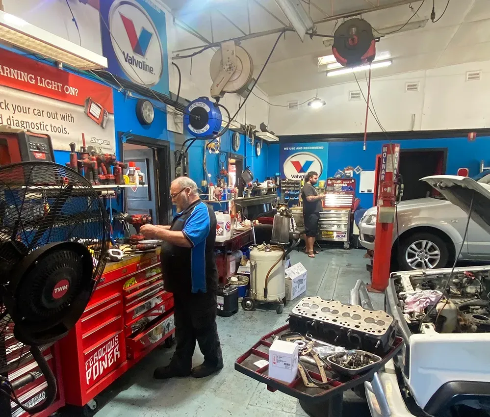 Mechanic working in a busy auto repair shop with tool chests, car parts, and a lifted vehicle — M T Automotive In Wallsend, NSW