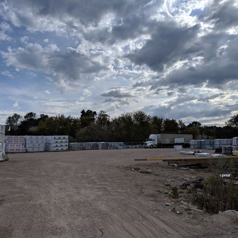 Construction materials yard under a cloudy sky with pallets, truck, and trees in the background.