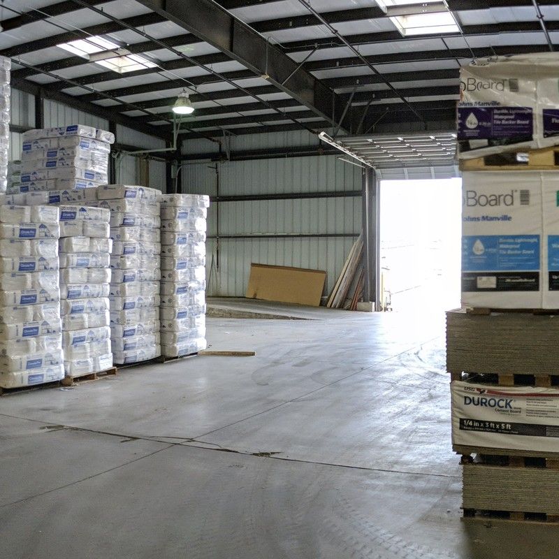 Inside of a warehouse. Pallets of building supplies stacked on the concrete floor. Sunlight through open doorway.