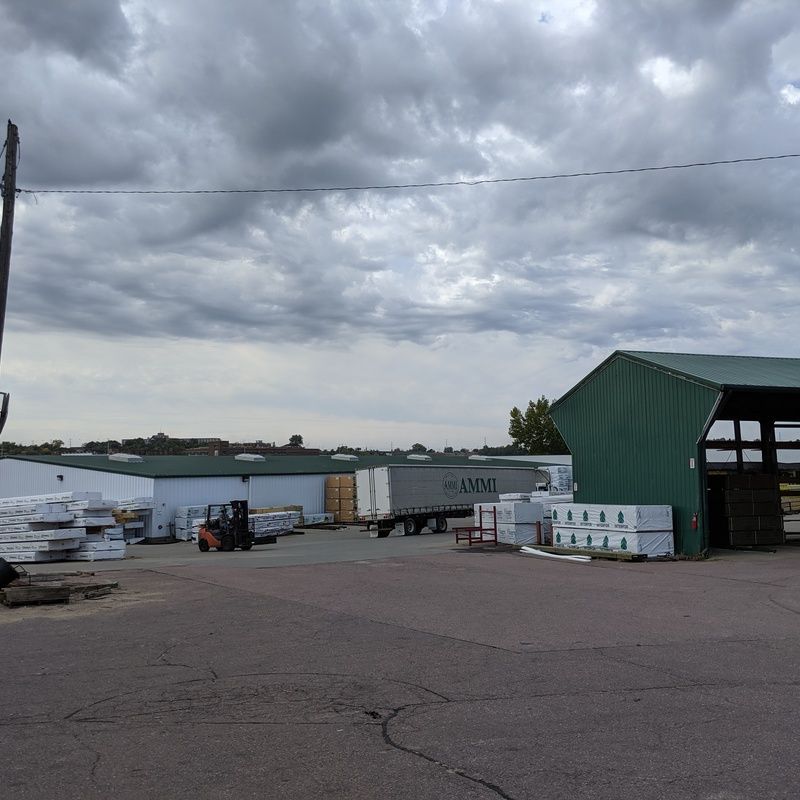 A lumber yard with a forklift, semi-truck, and green shed under a cloudy sky.