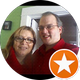 Two people smile at the camera. A woman with glasses and a man in a red shirt stand close together indoors.