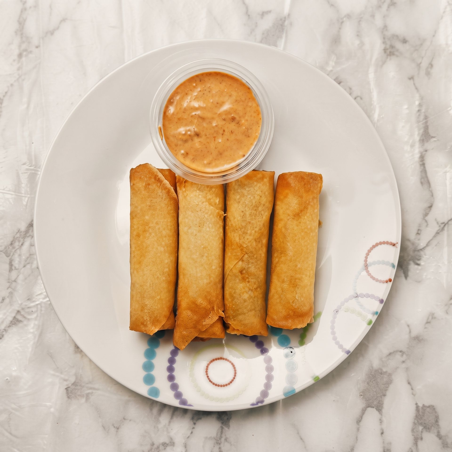 Four golden fried spring rolls on a white plate with a small cup of orange dipping sauce