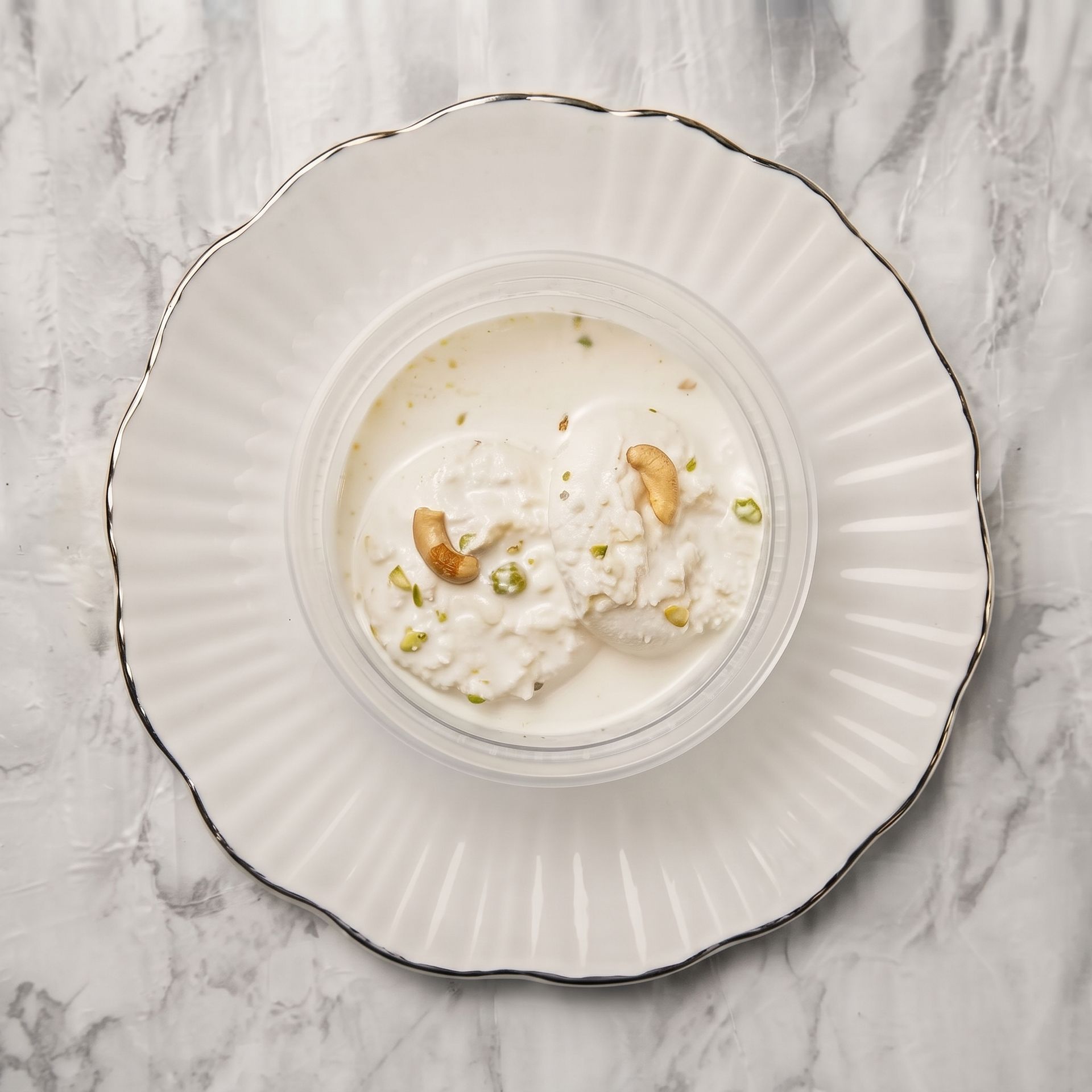 White dessert in a fluted bowl on a white marble table, topped with cashews and pistachios