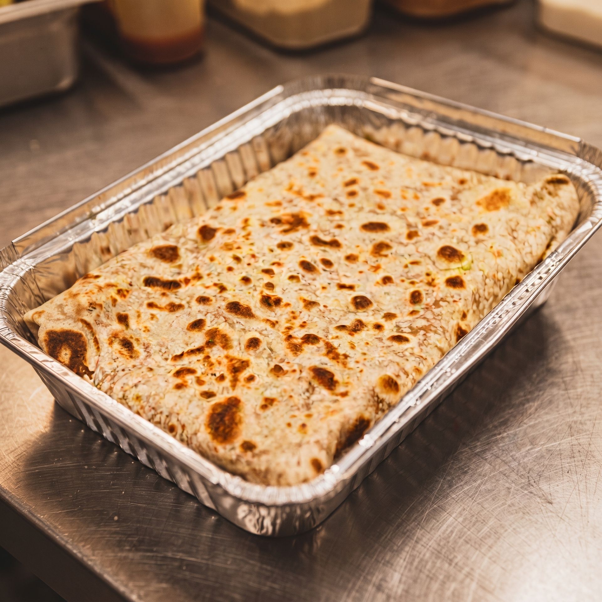 Foil tray of baked flatbread with browned spots on a kitchen counter