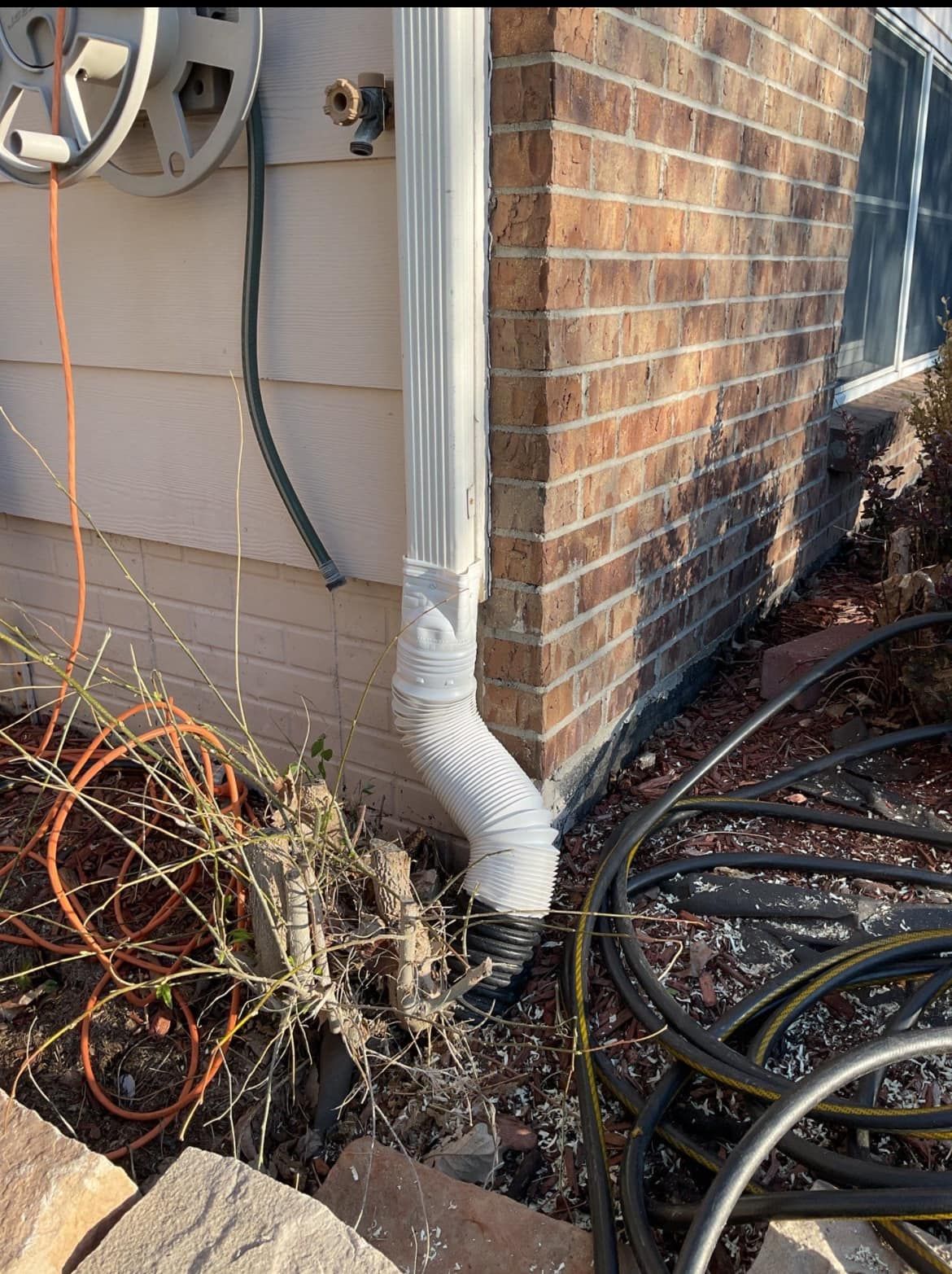 A drainpipe is sitting on the side of a brick house next to a bunch of hose.