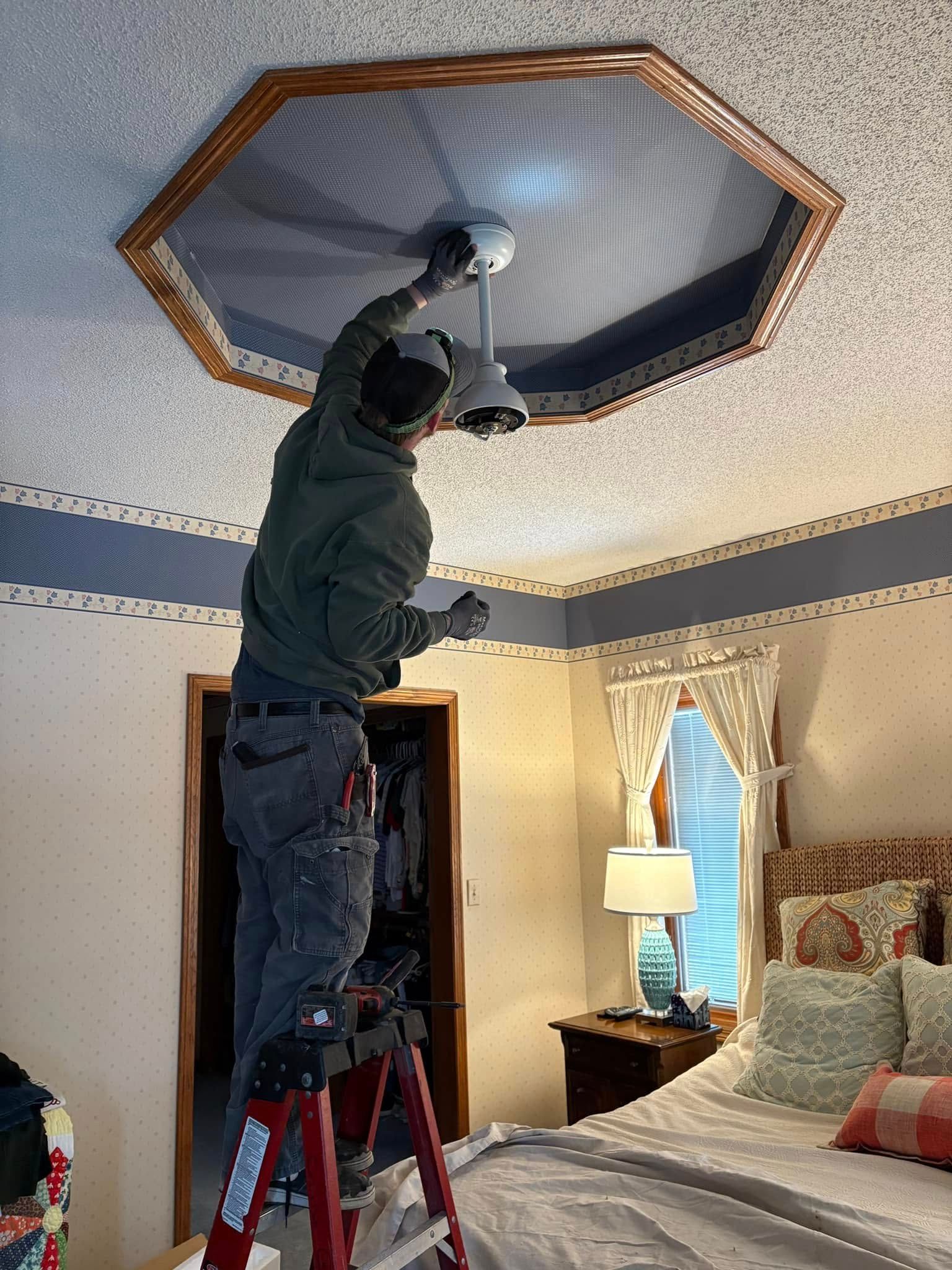 A man is standing on a ladder fixing a ceiling fan in a bedroom.