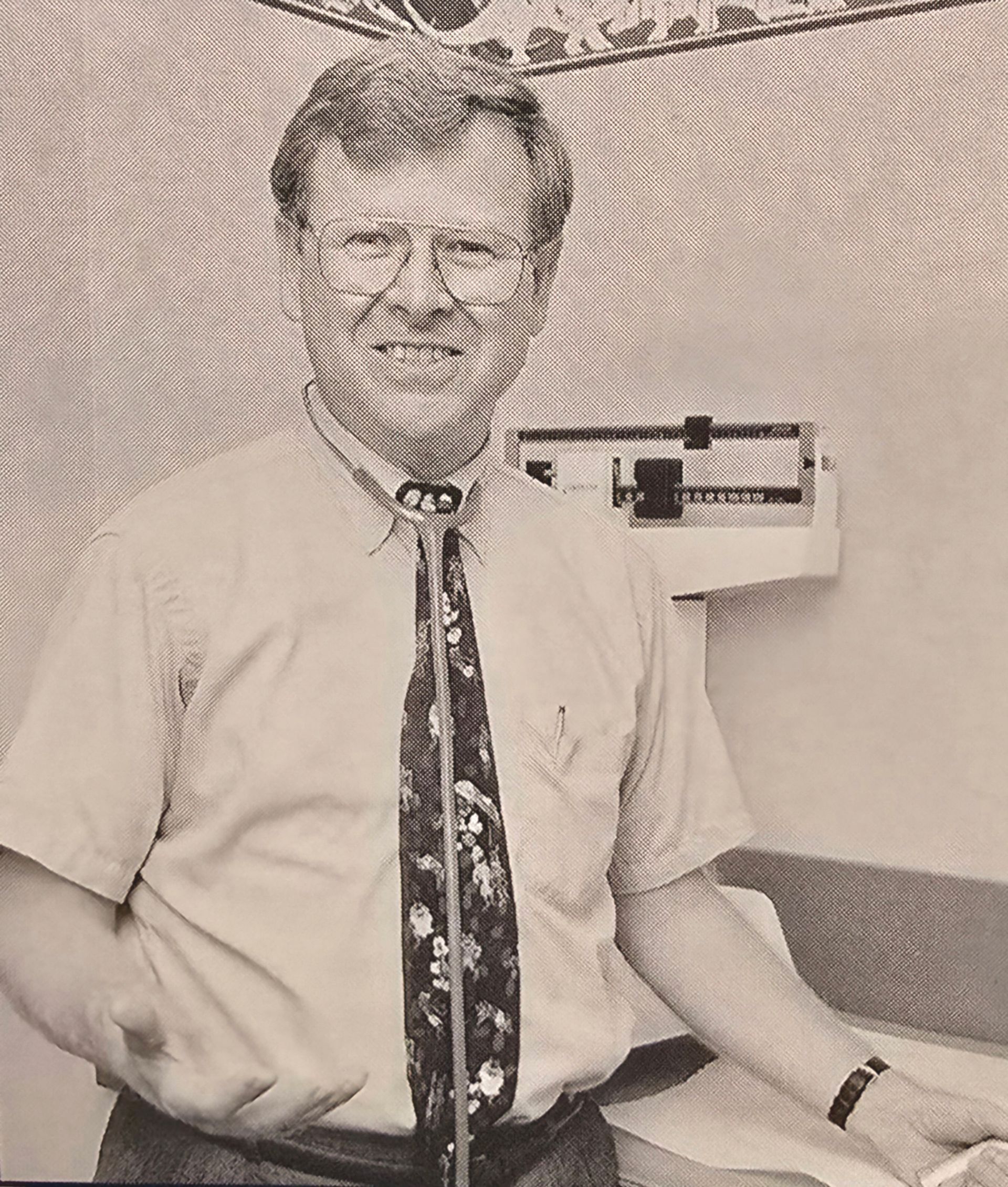 Man standing in a room, smiling, wearing a white shirt and patterned tie near a wall-mounted device.