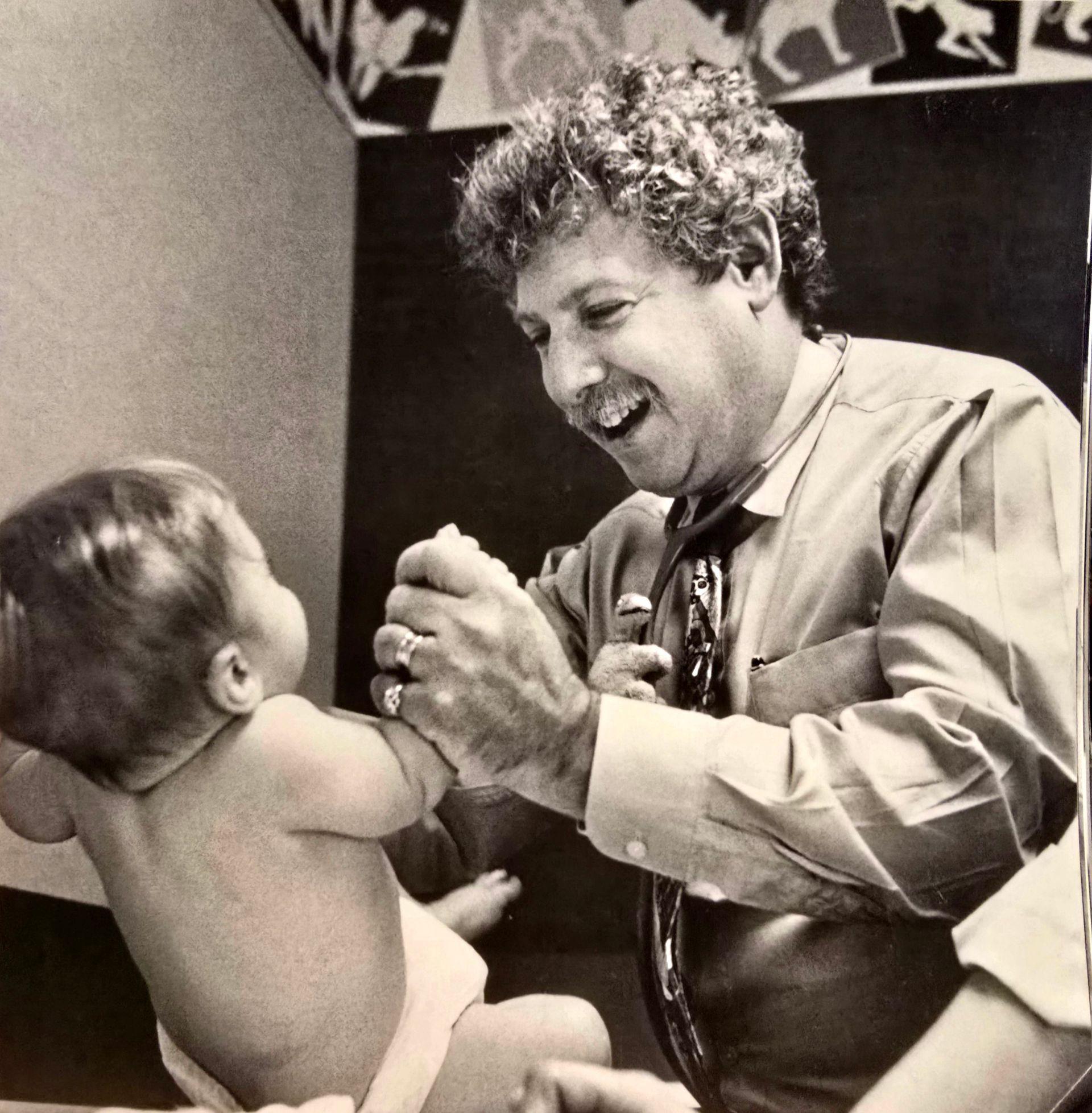 Smiling adult holding hands with a baby in a diaper, facing each other indoors.
