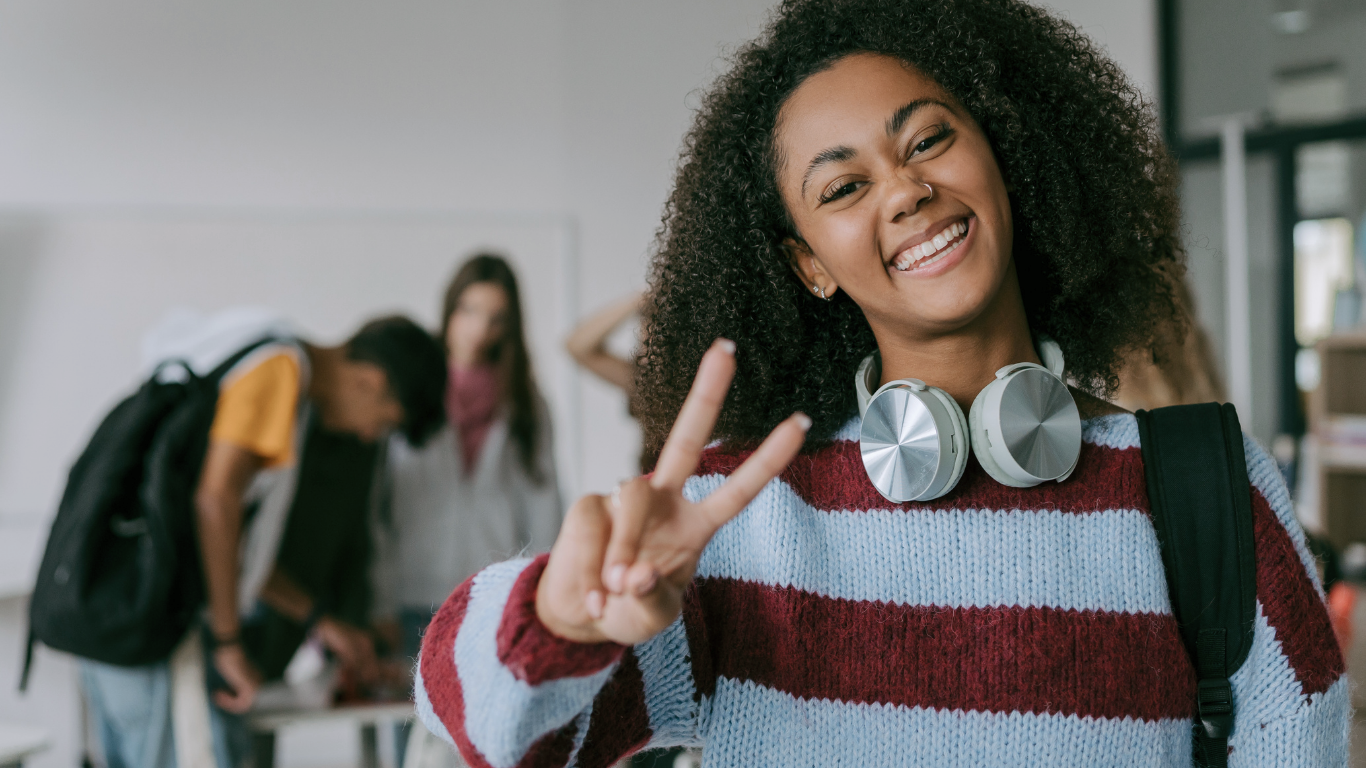 Smiling student in a striped sweater makes a peace sign in a hallway, with headphones around her neck.