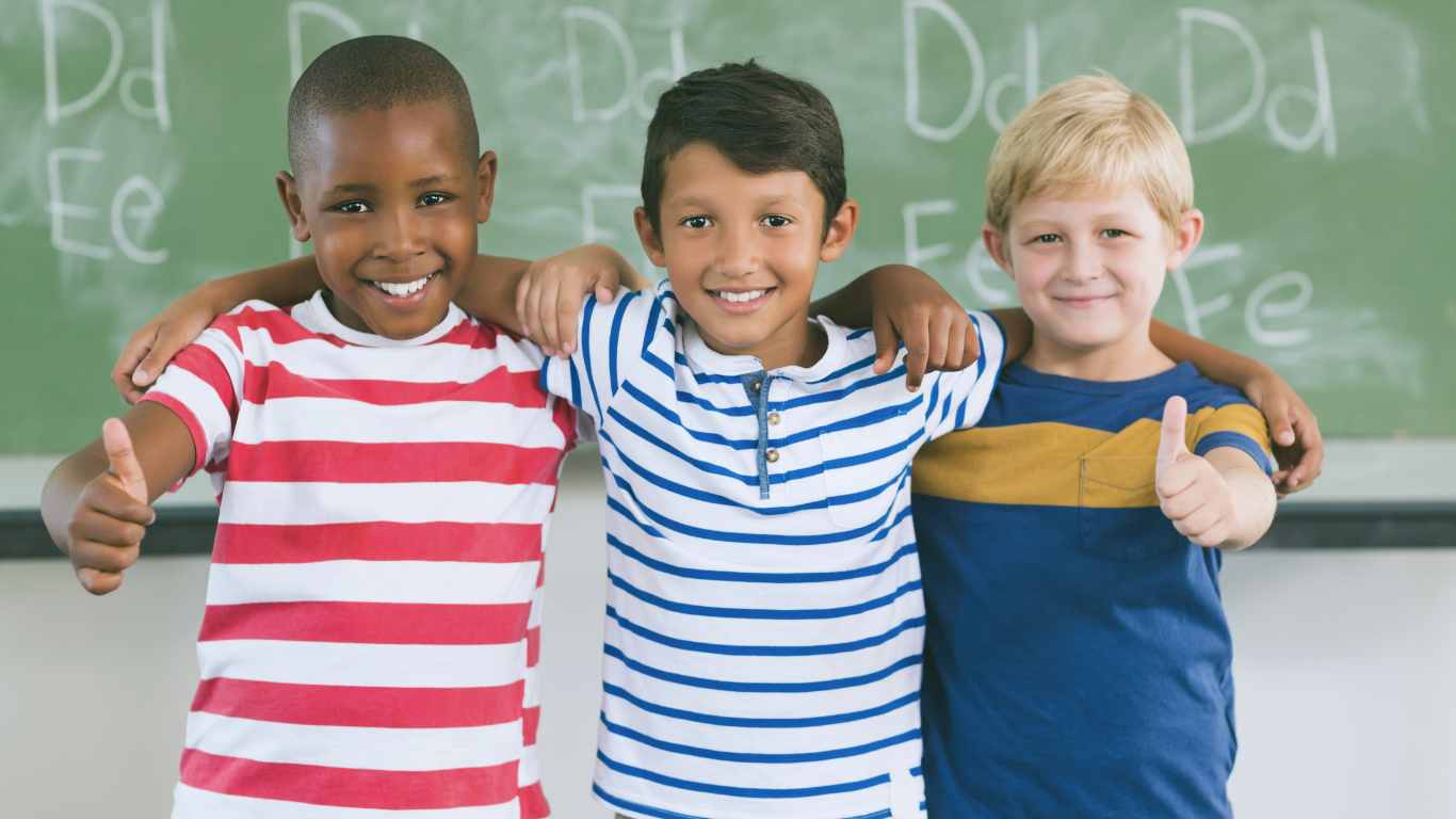 Three smiling children standing arm in arm in front of a chalkboard, each wearing a striped shirt