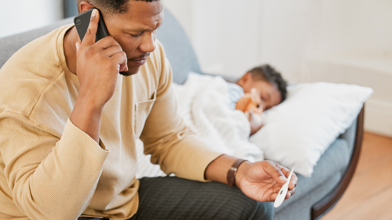 Man sitting on a bed, talking on a phone while checking a tablet, with a child resting behind him.