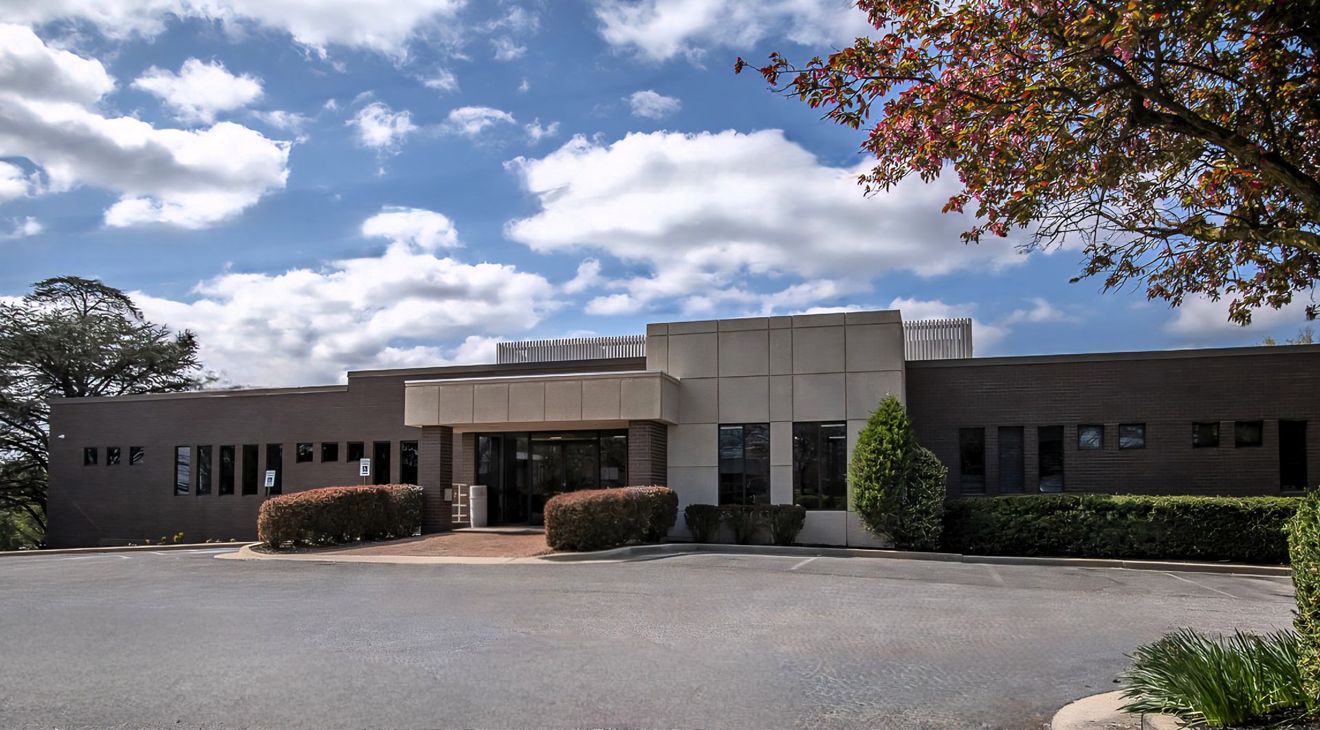 Single-story office building with a glass entrance, gravel driveway, and cloudy sky overhead