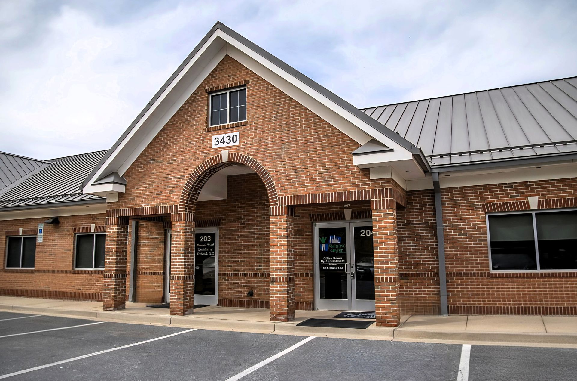 Brick church building entrance with arched doorway and gray metal roof, viewed from a parking lot