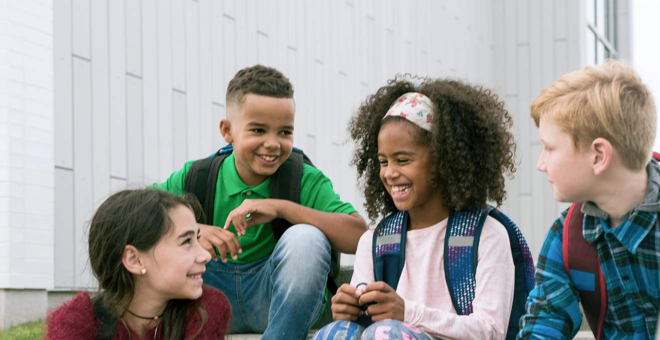 Four children sit outdoors on steps, smiling and talking together, in casual colorful clothes.