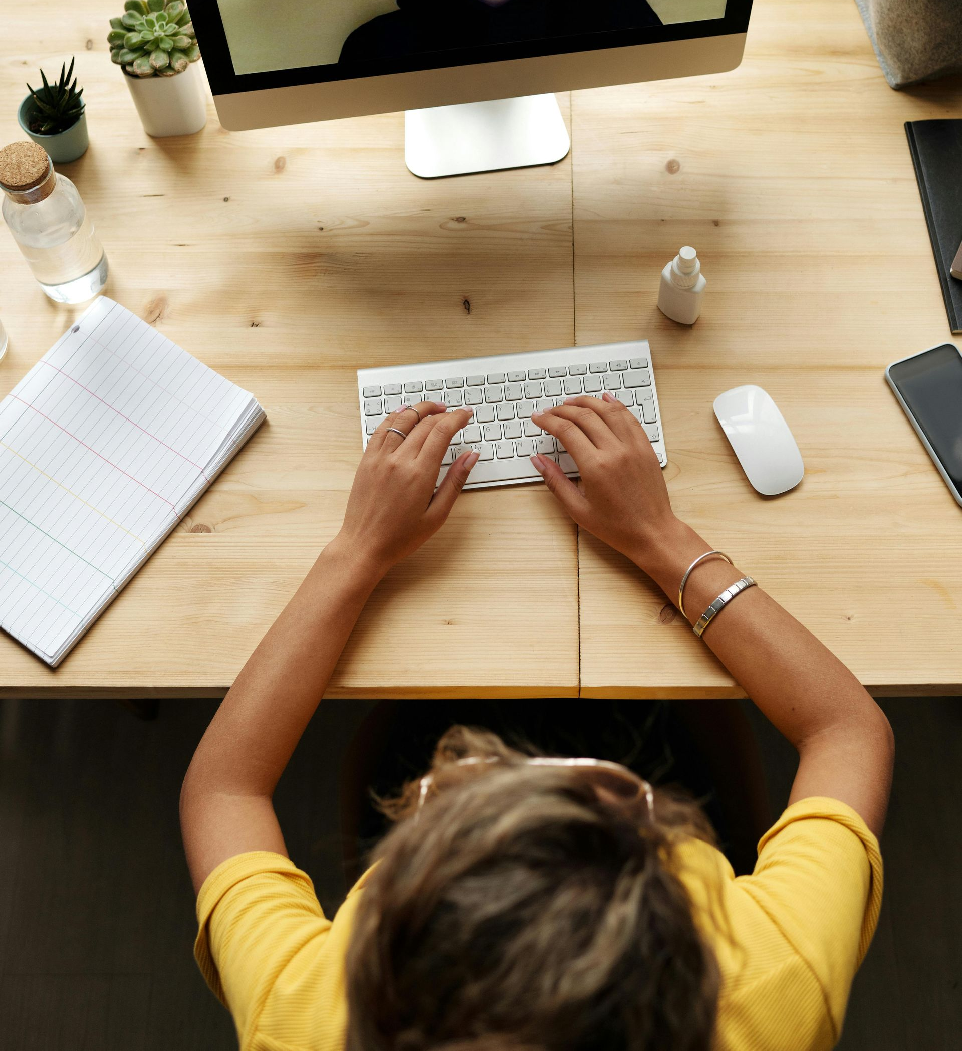 Person typing at a white keyboard on a wooden desk with a monitor and mouse nearby