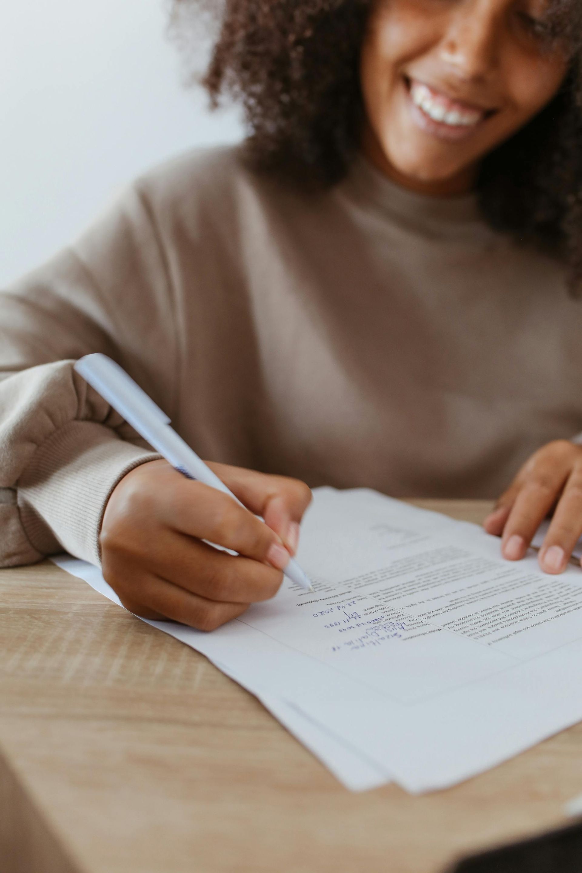 Person writing on a document with a pen at a wooden desk, smiling.
