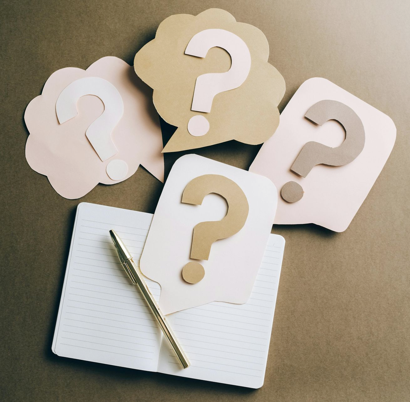 Question-mark cutouts on notepads with a pen, arranged on a brown background.