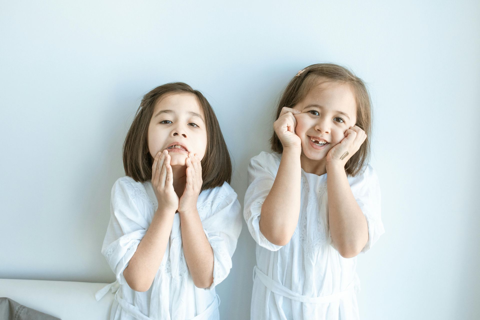 Two children in white dresses with hands on cheeks, smiling against a pale blue wall.
