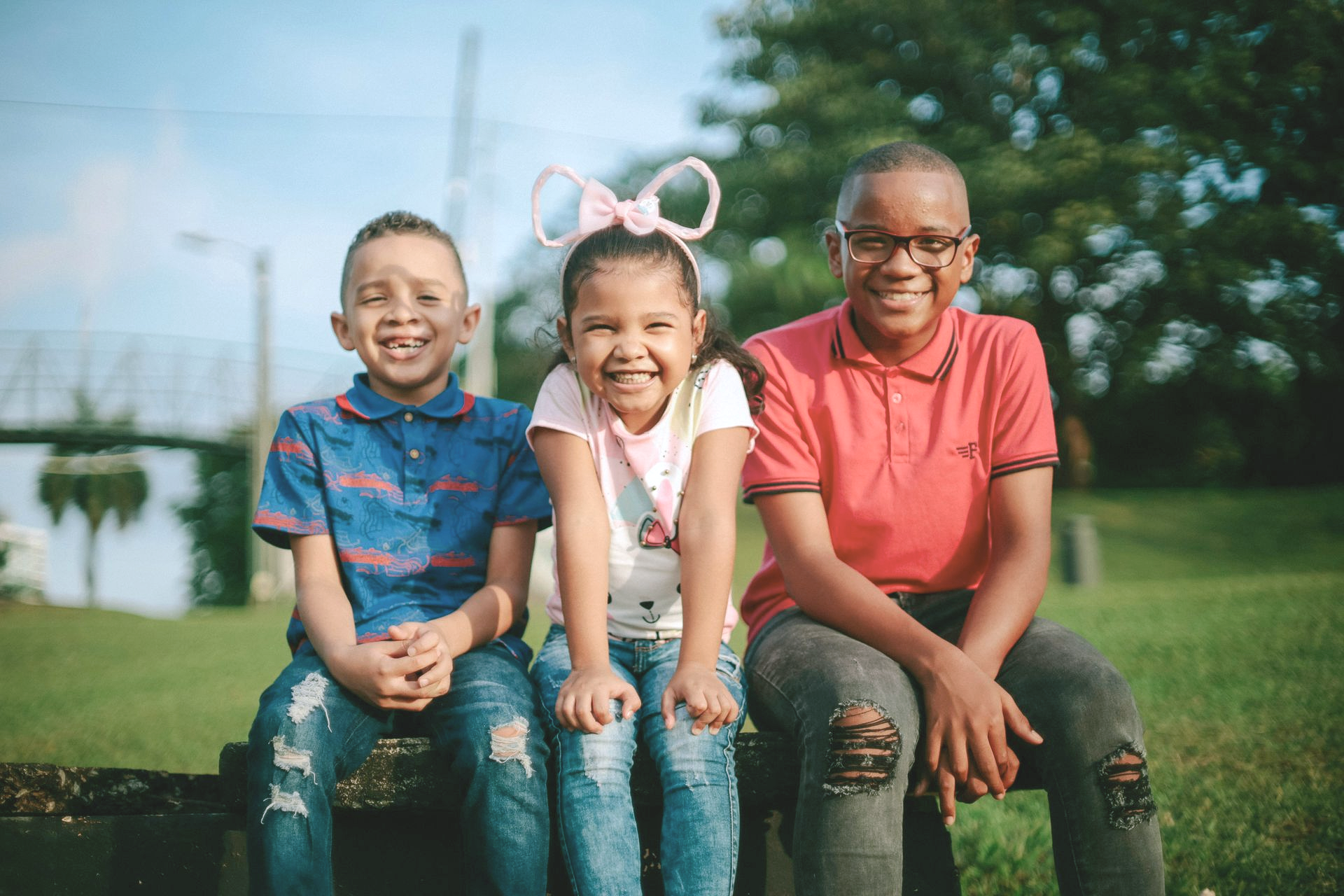 Three children sitting on a bench outdoors, smiling in a sunny park.