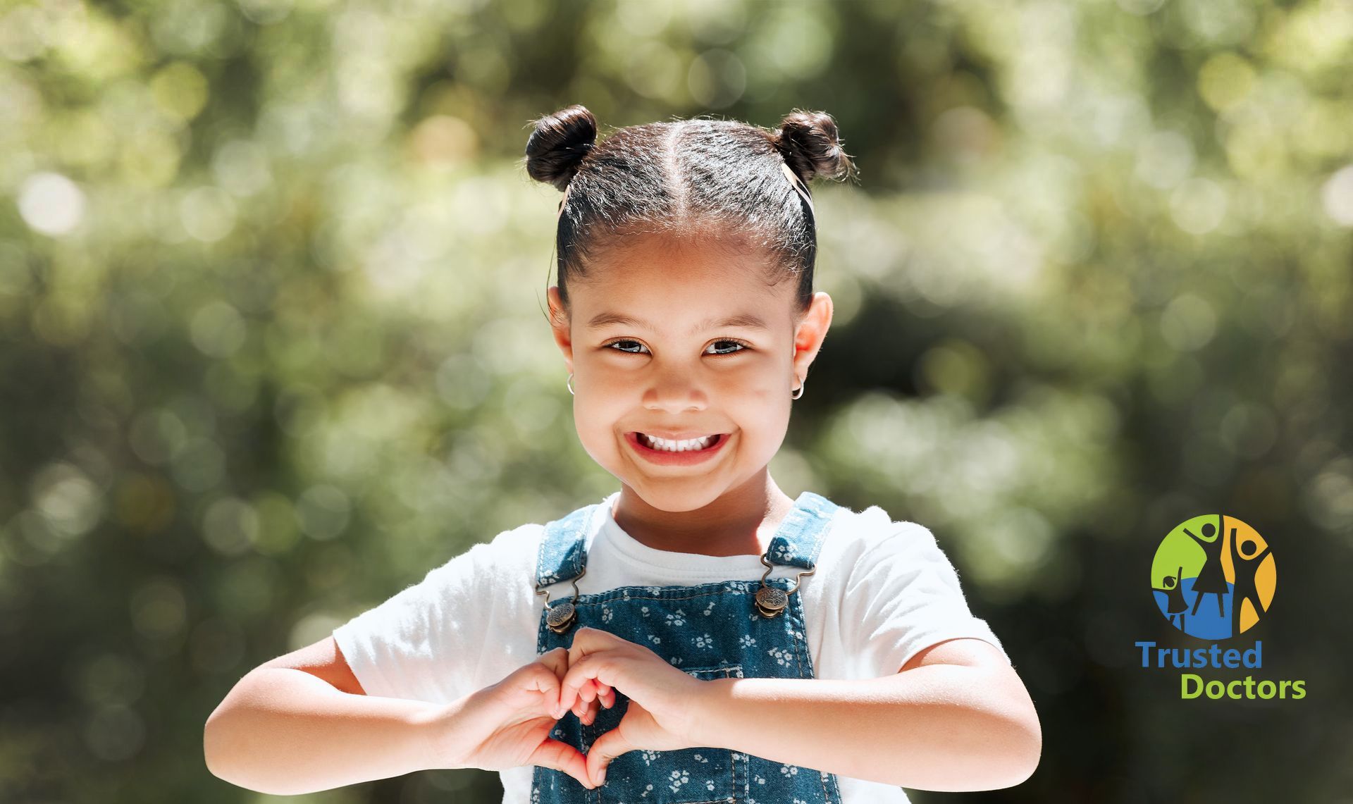 Smiling child with braided hair and blue overalls making a heart shape with hands outdoors
