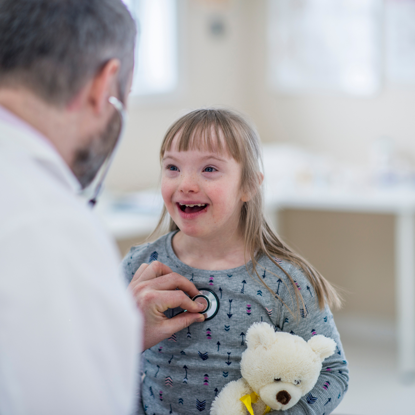 Doctor examining smiling child holding a teddy bear in a bright clinic room