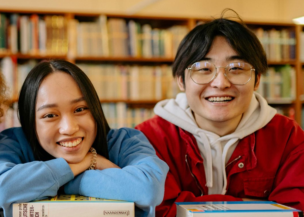 Two smiling students in a library, leaning on books with shelves in the background