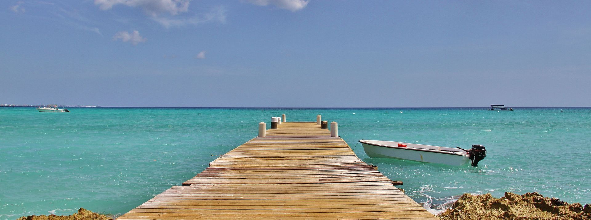 Wooden pier extends into turquoise water; small boat tied nearby, blue sky overhead.