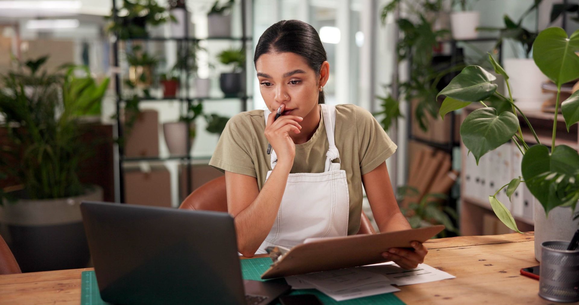 Person studying at a wooden table with a laptop, notebook, and plants in a bright home office