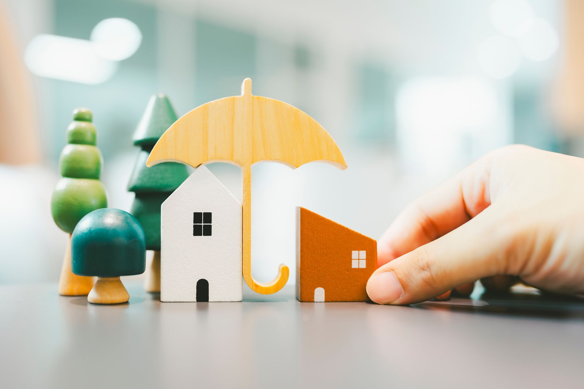 Hand placing a small orange house beside a white house under a yellow umbrella with toy trees in the background