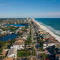 Aerial view of a coastal town with a beach, ocean, road, and homes under a clear blue sky.