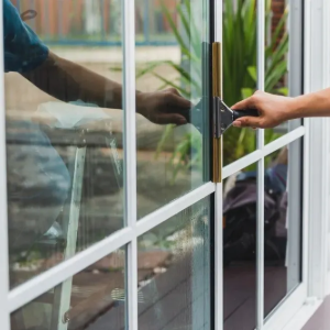 Two hands opening a glass door, with reflections and greenery visible outside