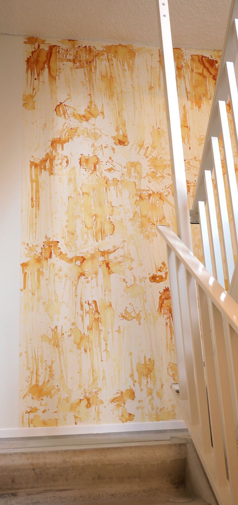 Een muur in een trappenhuis met een abstracte, gedruppelde textuur van gele en oranje verf op een witte ondergrond.
