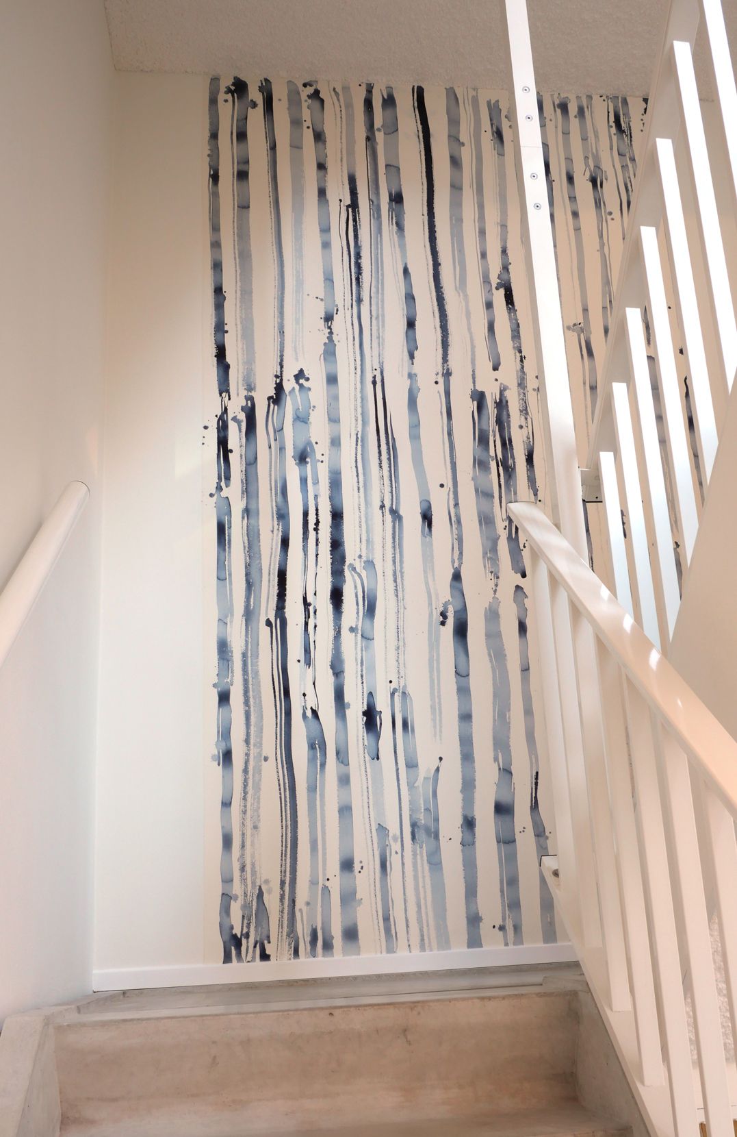 Een trappenhuiswand met een verticaal, abstract behangpatroon van blauwe en grijze penseelstreken op een witte achtergrond.