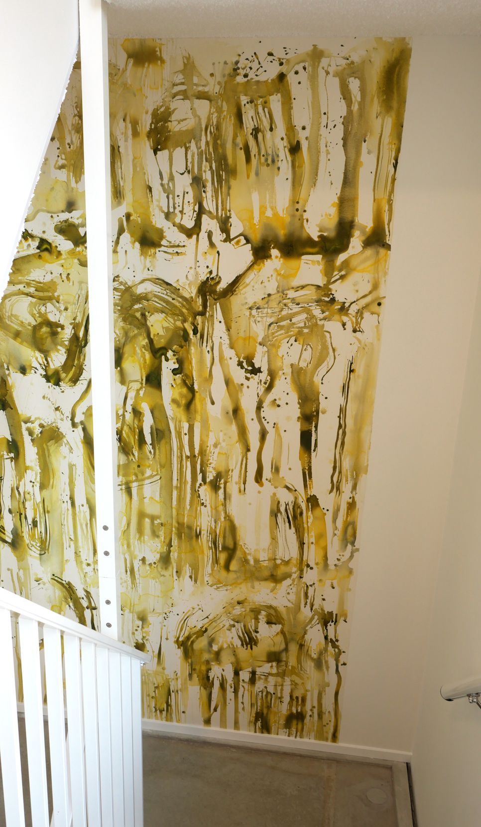 Een verticaal abstract muurschildering met geelgroene waterverfdruppels en -spatten op een witte trappenmuur.